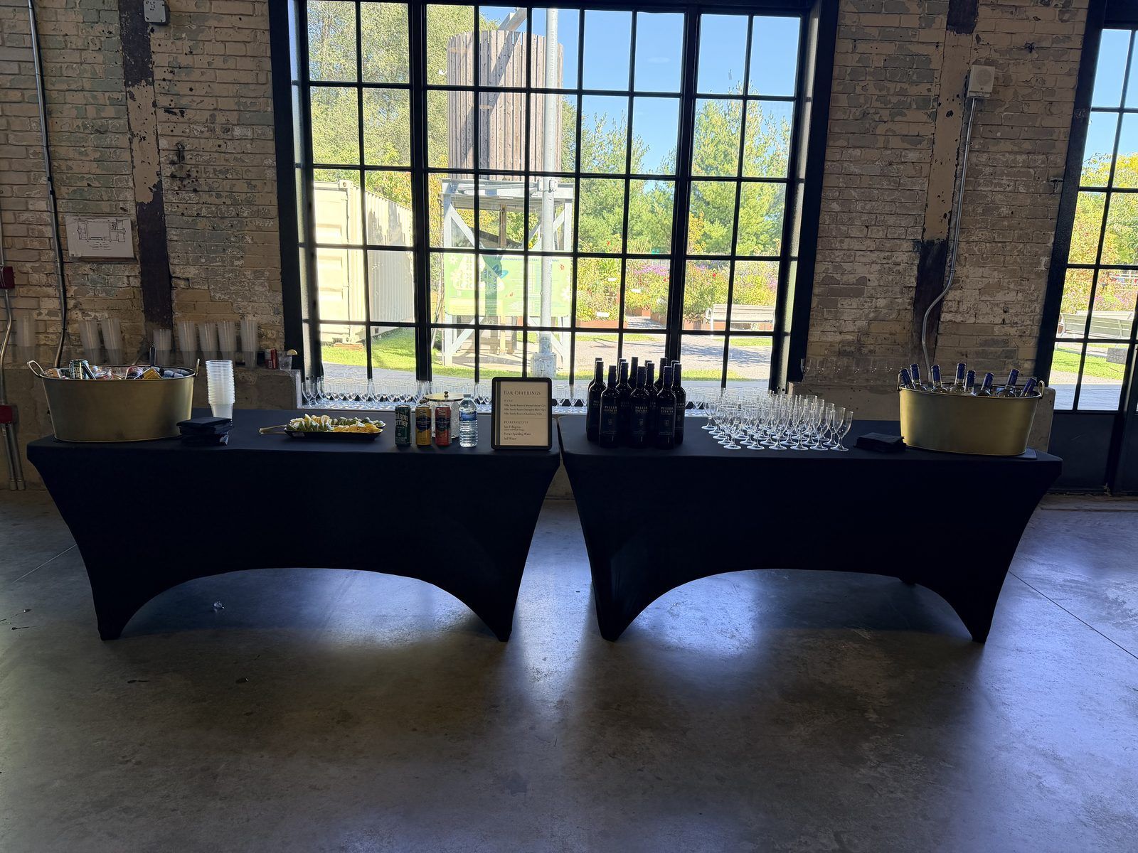 Two bar tables set with glassware in front of tall warehouse windows