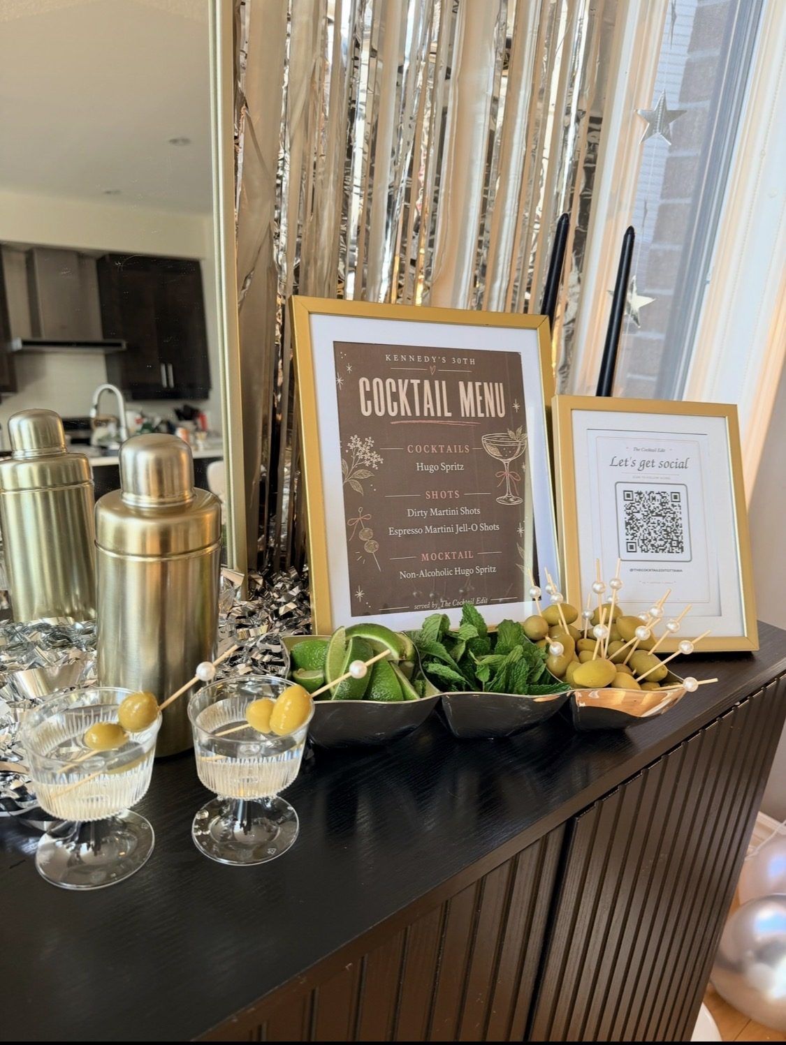 A cocktail menu, garnishes and a shaker on a dark bar top